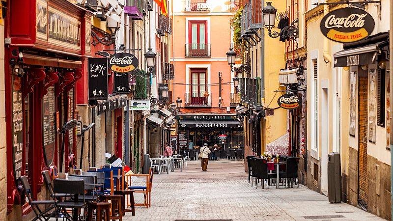 Calle española con fachadas coloridas, terrazas y carteles de "Coca-Cola" y "MAJADERITOS". Una persona camina de espaldas hacia el fondo.