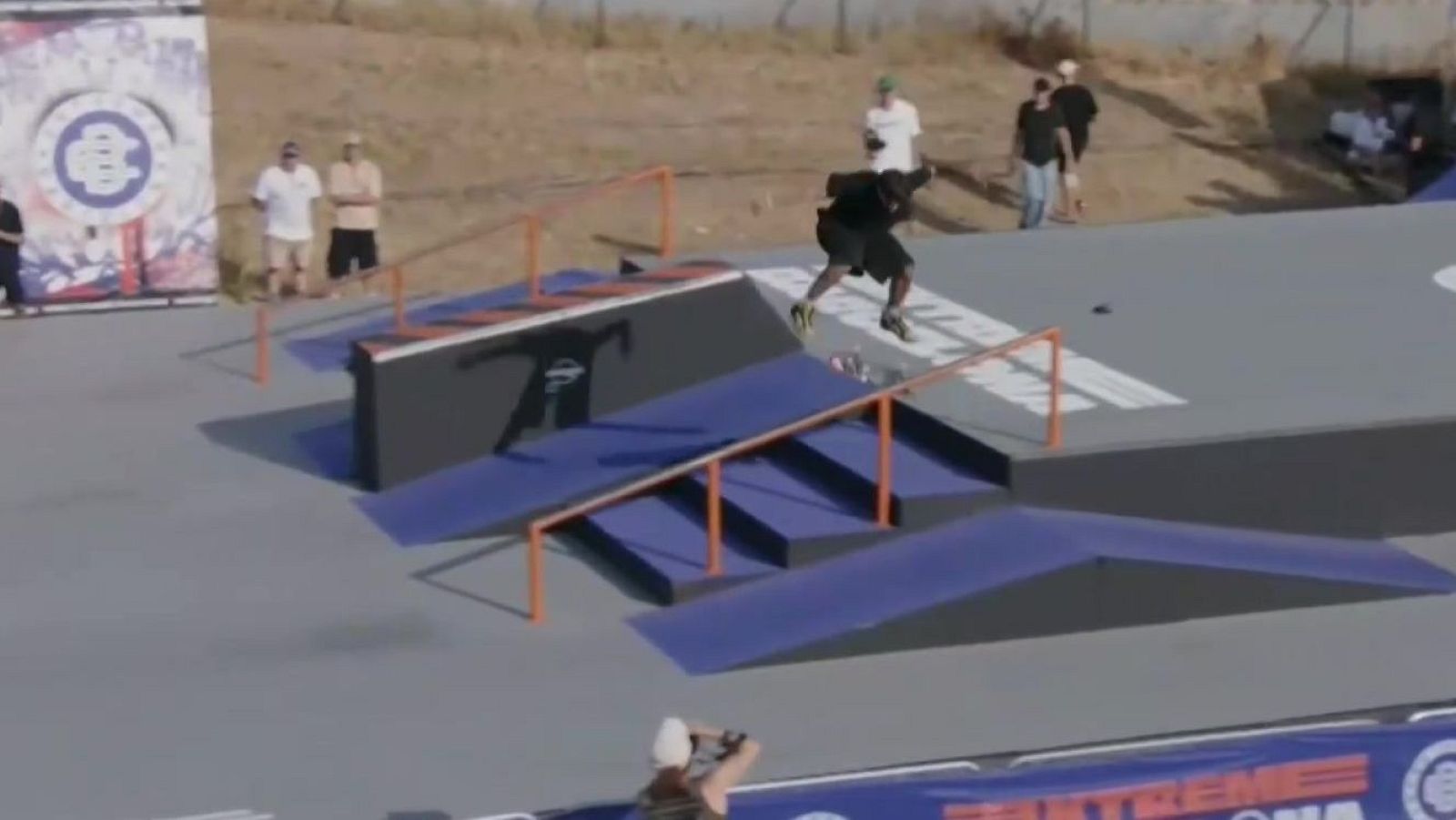 Joven patinador en pleno salto acrobático en un skatepark, con estructuras azules y grises, barandillas naranjas y un banner "EXTREME" al frente. Fotógrafo en primer plano.