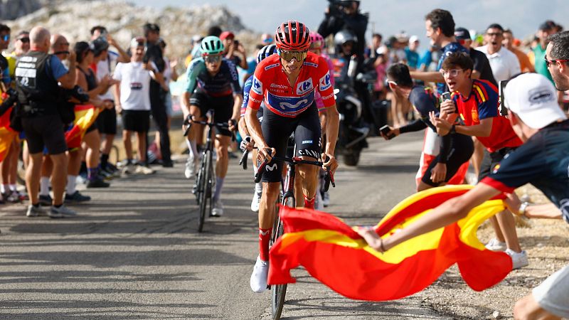 Enric Mas del equipo Movistar, con maillot rojo, azul y blanco, pedalea intensamente en una carrera ciclista. El público anima con fervor, ondeando una bandera de España.