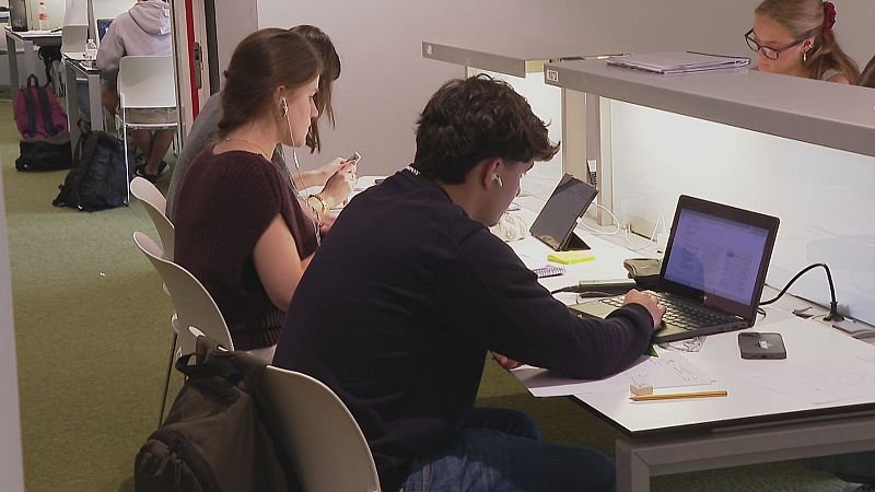 Jóvenes estudian concentrados en una sala moderna con escritorios individuales, portátiles, móviles y otros materiales de estudio. Ambiente académico y de preparación para exámenes.