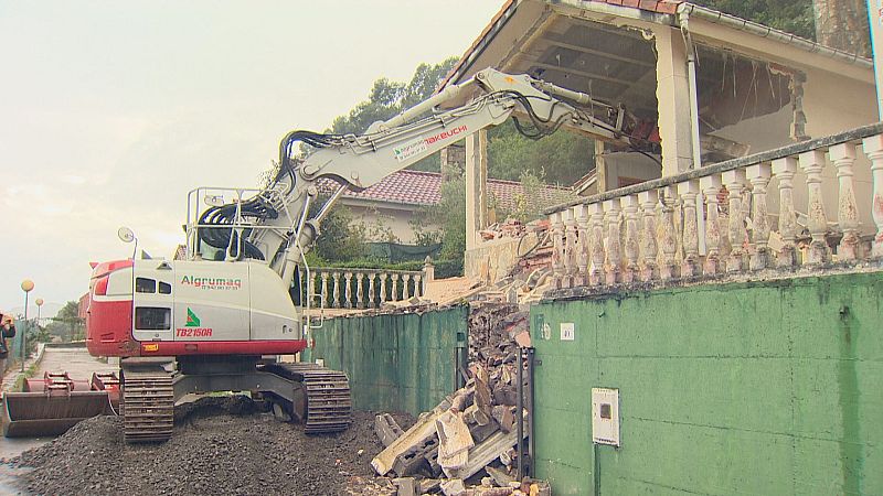 Excavadora "Algrumag TB2150R" derriba vivienda en Argoños. Se aprecian tejado rojizo, barandilla dañada y abundantes escombros. Fondo de vegetación y edificaciones.