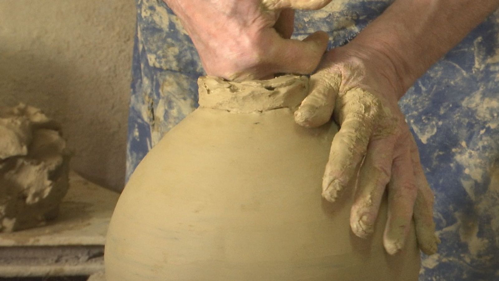 Manos con arcilla moldean una vasija de barro, trabajando el cuello de la pieza. Se aprecian detalles de la arcilla y la prenda azul del artesano.