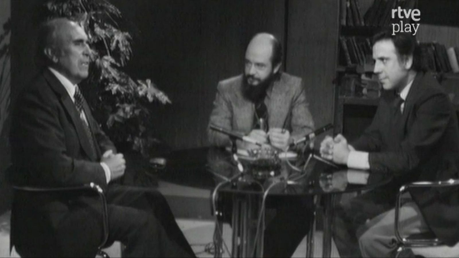 Fernando Jiménez del Oso, a la derecha, con traje oscuro, participa en una tertulia en el plató de "Más allá" junto a dos invitados. La escena, en blanco y negro, muestra micrófonos sobre la mesa y un fondo con libros.