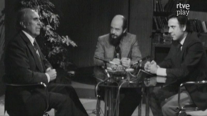 Fernando Jiménez del Oso, a la derecha, con traje oscuro, participa en una tertulia en el plató de "Más allá" junto a dos invitados. La escena, en blanco y negro, muestra micrófonos sobre la mesa y un fondo con libros.
