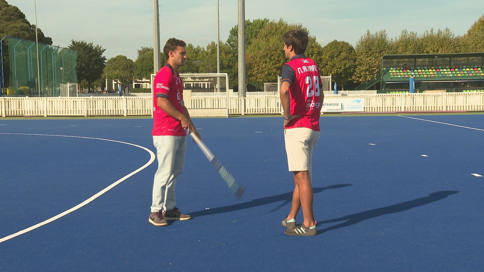 Jugadores de hockey hierba en campo azul. Uno luce "N. ALARPE 28" en su camiseta. Sostienen palos y posan para una entrevista, con vallas y gradas al fondo.