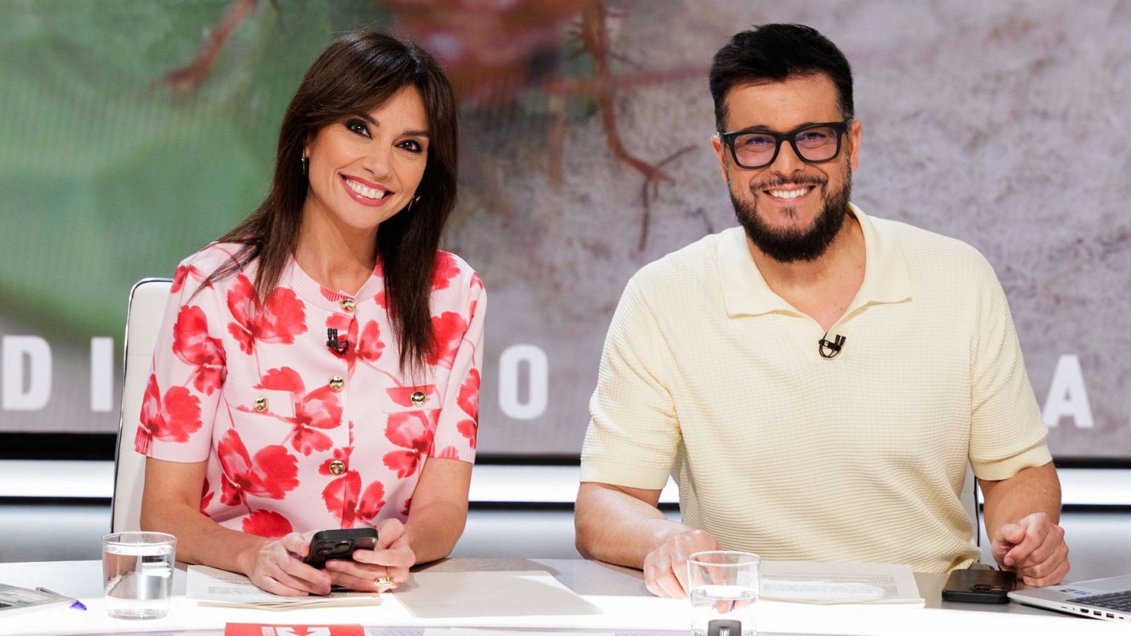 Nuria Marín y Omar Suárez, presentadores, sonríen en un plató. Nuria con top rosa floral, Omar con polo amarillo y micrófono. Pantalla con "DI" y "O" al fondo.