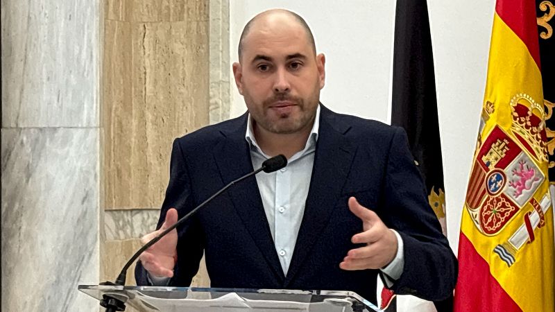 Primer plano de Miguel Ángel Pérez Triano, Delegado de Gobierno en Ceuta, en rueda de prensa. Hombre calvo con barba, traje azul y camisa clara, gesticula mientras habla. Se ven banderas de España y otra oscura, y paredes de mármol.