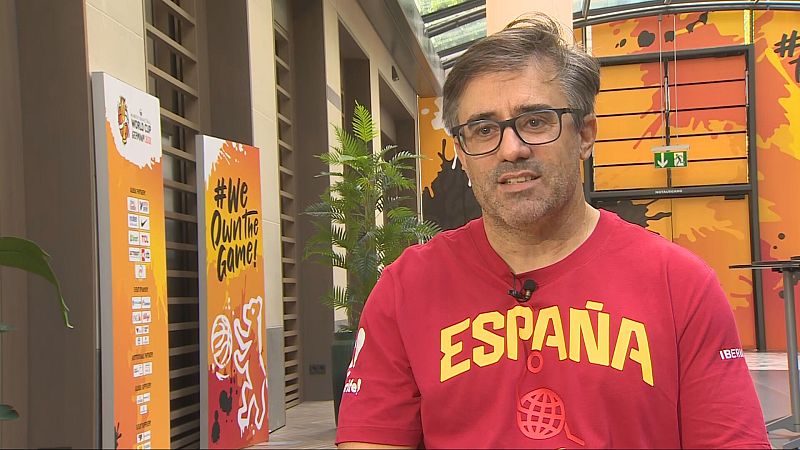 Periodista deportivo español Oscar Viana con camiseta "ESPAÑA" e "IBERIA", gafas y barba, frente a paneles de evento deportivo con "#We Own The Game!".