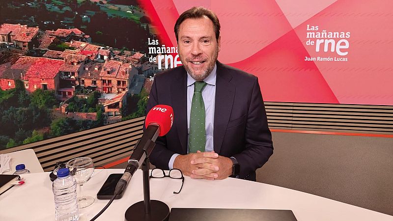 Óscar Puente en estudio de radio, sonriente, con traje oscuro, camisa azul y corbata verde. Fondo con "Las mañanas de rne" y "Juan Ramón Lucas".