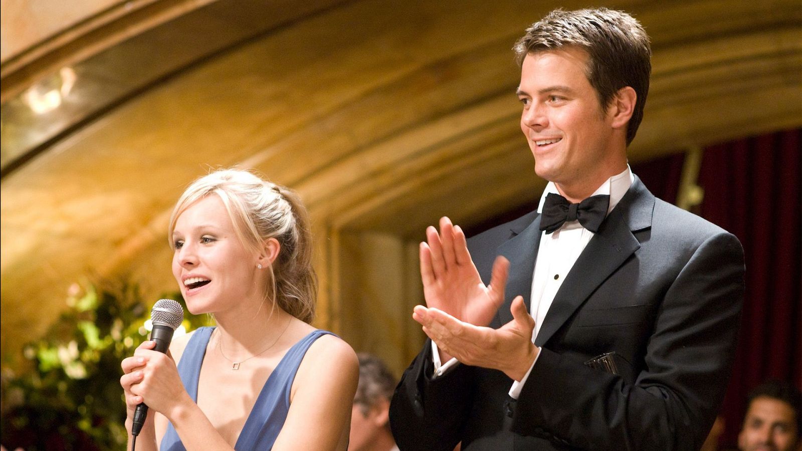 Kristen Bell habla al micrófono con entusiasmo, mientras Josh Duhamel aplaude en un evento formal. Ambos actores lucen elegantes en un salón decorado.