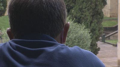 Hombre de espaldas, vestido con polo azul oscuro, contempla un paisaje exterior con vegetaci�n y arquitectura de piedra. Sugiere introspecci�n.
