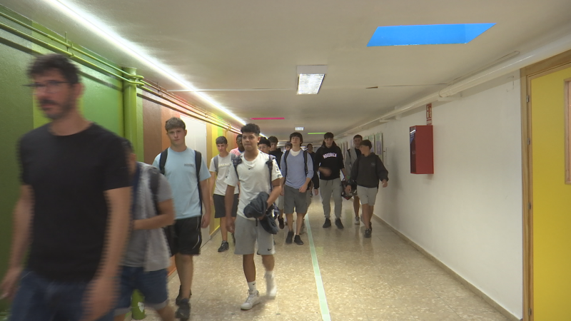 Pasillo institucional con hombre con gafas y grupo de jóvenes en movimiento. Luces fluorescentes, paneles verdes y blancos, extintor rojo y puerta amarilla.