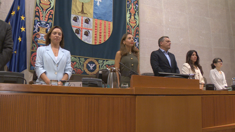 Cinco políticos, incluyendo a Marta Gastón, Mar Vaquero, Arturo Aliaga, Ana Marín e Isabel Lasobras, posan de pie en un estrado durante una sesión plenaria en las Cortes de Aragón, con el escudo de Aragón y la bandera de la UE al fondo.