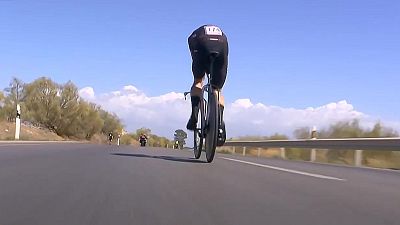 Ciclista con dorsal 174 en contrarreloj, postura aerodin�mica, carretera soleada con vegetaci�n seca y veh�culos de apoyo al fondo.