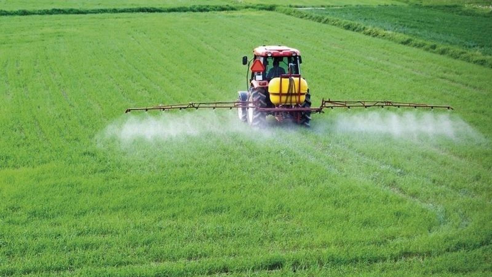 Tractor rojo con pulverizador y tanque amarillo trabajando en un campo verde, liberando rocío sobre los cultivos bajo luz solar.