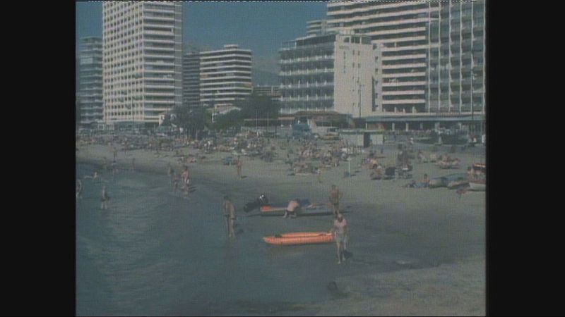 Vista panorámica de una playa mediterránea concurrida en un día soleado, con bañistas en la arena y el mar, y edificios modernos al fondo.