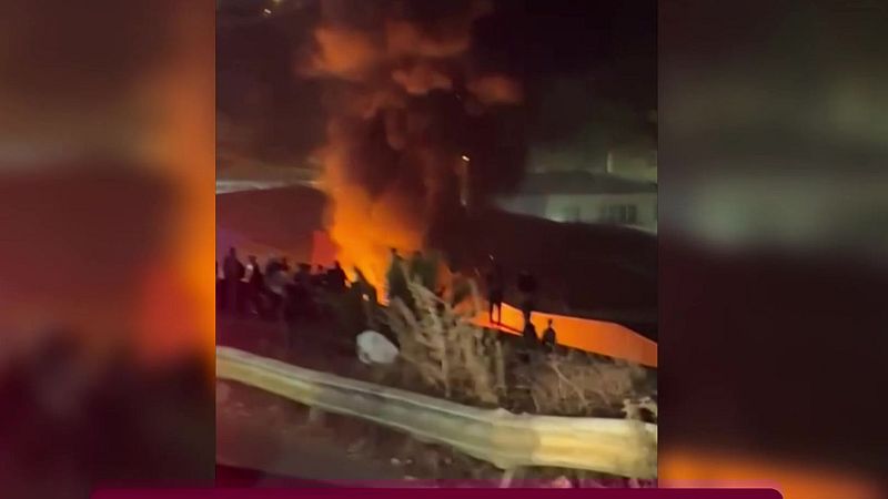 Gran incendio nocturno con llamas intensas y humo oscuro, observado por siluetas humanas. Se aprecian estructuras en primer plano y al fondo.