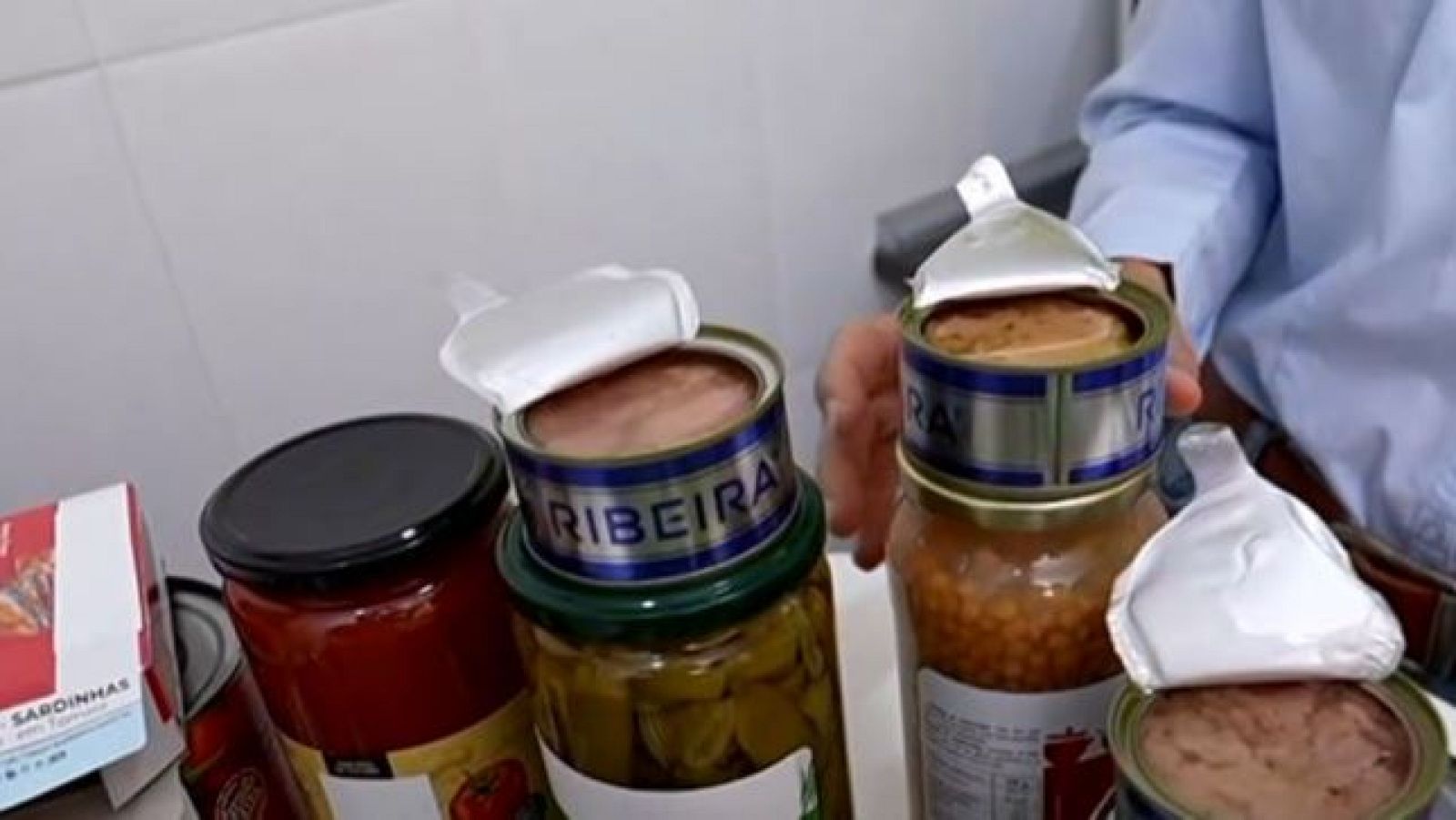 Primer plano de productos alimenticios envasados: latas abiertas de "RIBEIRA" con paté, tarros de salsa de tomate, encurtidos y sardinas. Fondo de azulejos blancos.