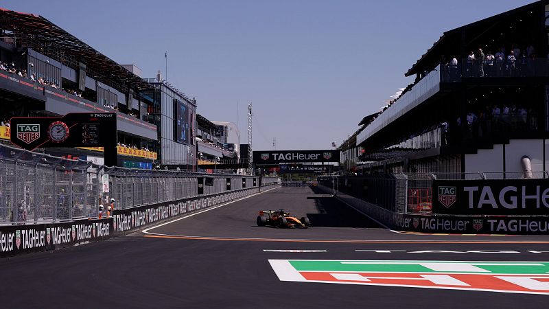 Coche de F1 naranja en circuito con gradas llenas y patrocinio de TAG Heuer, incluyendo reloj y pantallas.