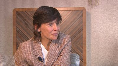 Periodista Ana Pastor, sentada, viste americana de cuadros y camiseta blanca. Micrófono de solapa visible. Fondo de madera y pared clara.