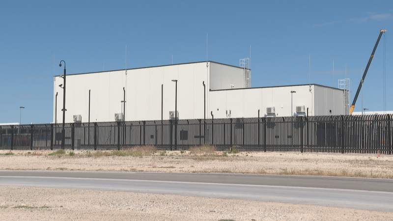 Edificio blanco moderno de aspecto industrial con valla de seguridad, cámaras y grúa de construcción visible. Terreno árido y carretera en primer plano bajo un cielo azul.