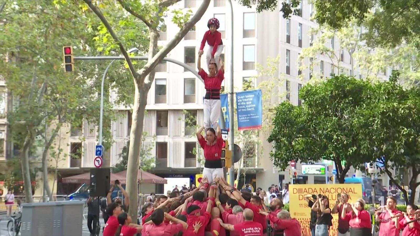Construcción de un 'castell' humano en la Diada Nacional de Catalunya, 11 de septiembre. Multitud observa, pancartas anuncian el evento. Personas vestidas de rojo forman la torre.