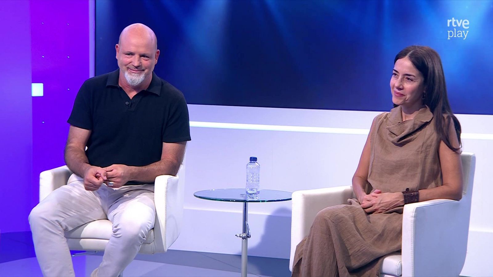 Juan Echanove y Aitana Sánchez-Gijón conversan en un plató de "rtve play", con fondo digital y una botella de agua sobre una mesa. Ambos visten ropa elegante y sonríen.