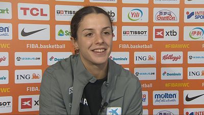 Elena Buenavida, jugadora de baloncesto espa�ola, sonr�e con chaqueta deportiva gris, logos de Nike y CaixaBank, y "ESPA�A" en el pecho. Fondo de patrocinadores con "FIBA.basketball" y "#FIBAWWC".