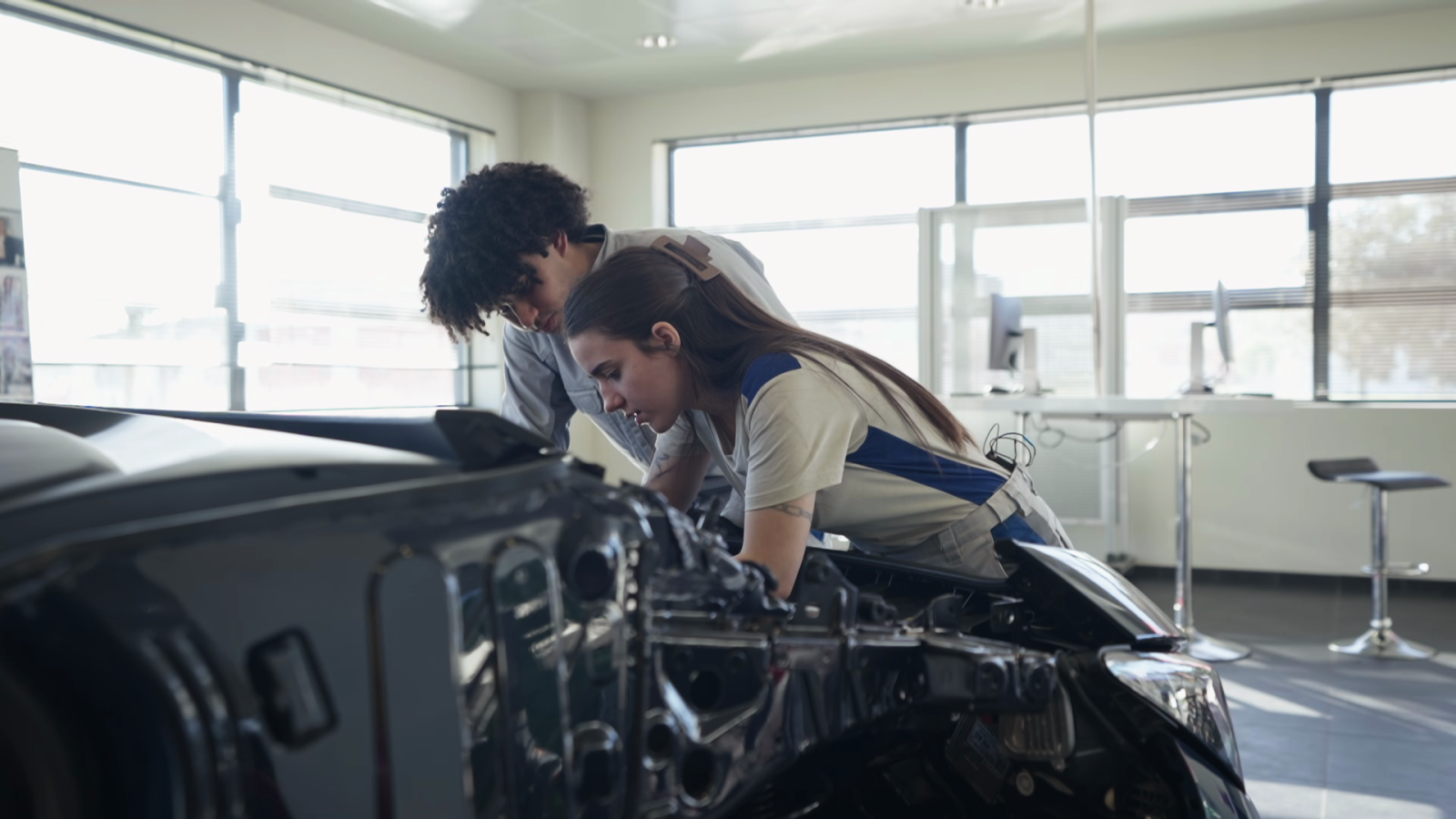 Jóvenes aprendices colaboran en la mecánica de un coche en un taller, con el motor expuesto y luz natural inundando el espacio.