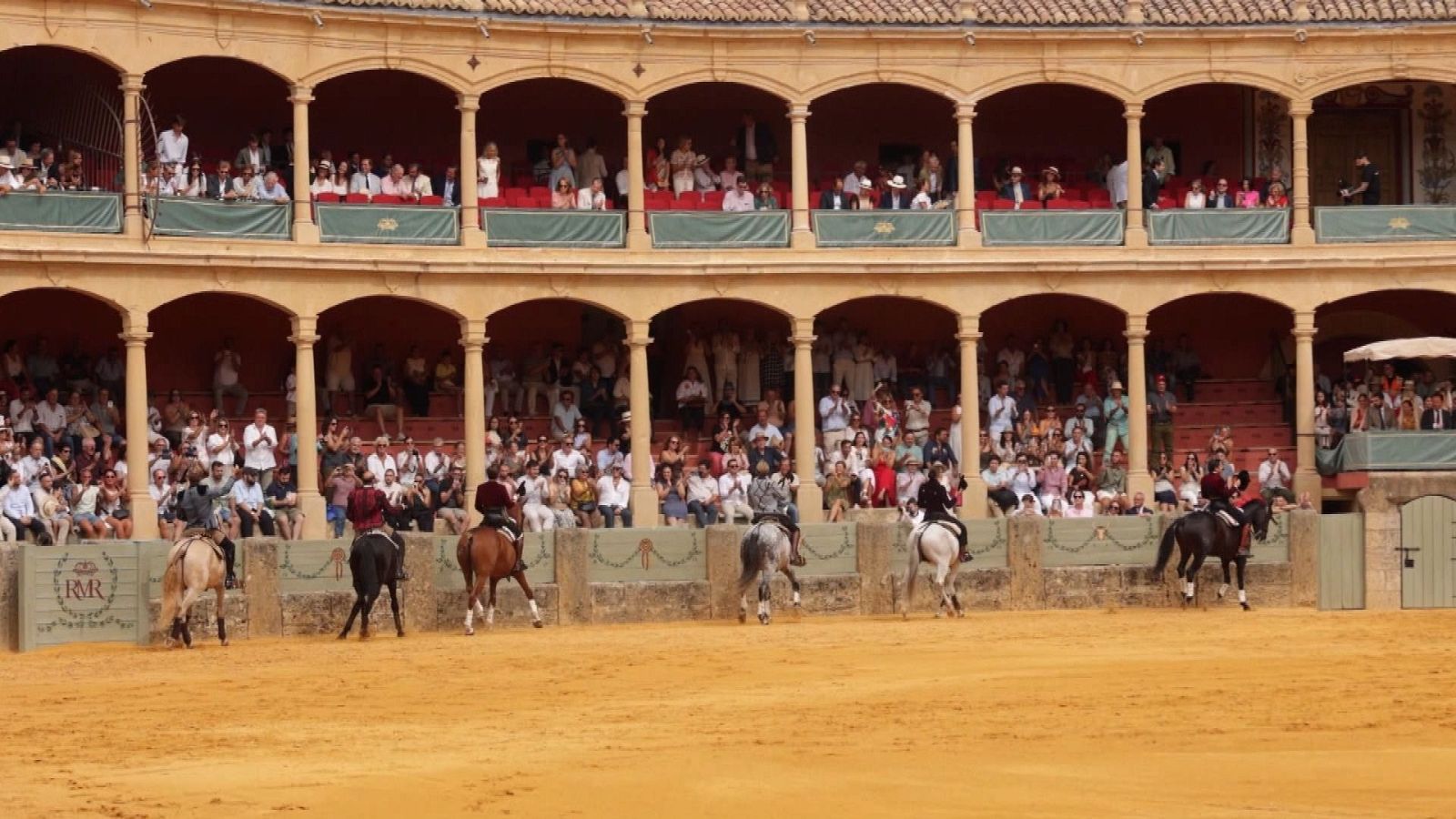 Plaza de toros con jinetes a caballo en la arena y público en los tendidos y palcos. Se observa el texto 'R&R' en la barrera.
