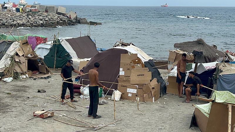 Asentamiento en playa con refugios de cartón y lonas. Cuatro hombres presentes. Mar y rompeolas al fondo.