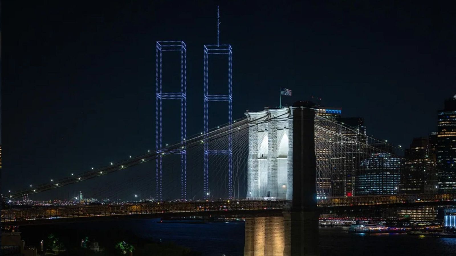 Vista nocturna de Nueva York con el Puente de Brooklyn y el memorial 'Tribute in Light', que evoca las Torres Gemelas, reflejados en el agua.