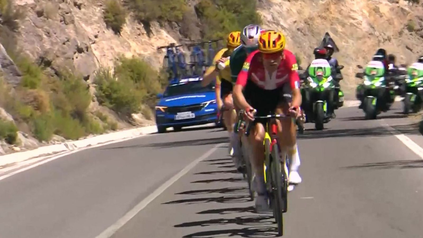 Ciclistas compiten en una carrera profesional en un día soleado, con un líder en maillot rojo y casco amarillo, escoltados por vehículos de apoyo en un paisaje montañoso.
