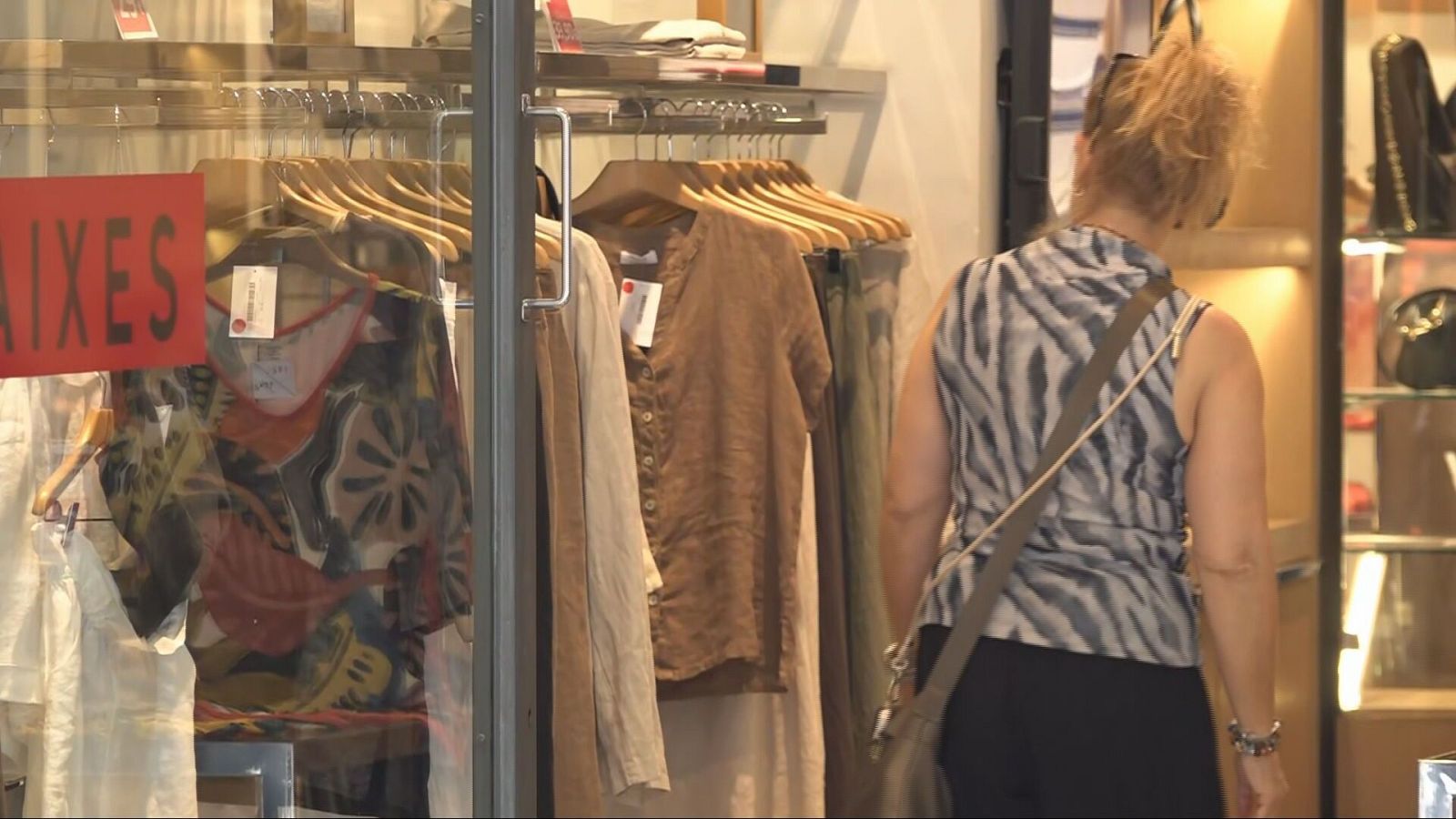 Mujer de espaldas con camiseta de cebra y bolso cruzado, frente a un escaparate con ropa y un cartel de "AIXES" (posiblemente "REBAIXES").