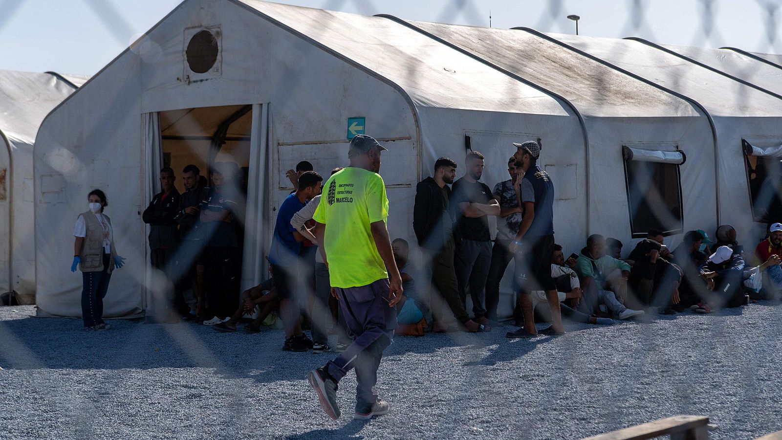 Campamento al aire libre con tiendas de lona, personal humanitario y personas en tránsito. Se observa una señal verde con flecha y un hombre con camiseta amarilla que dice "MARBELLO".