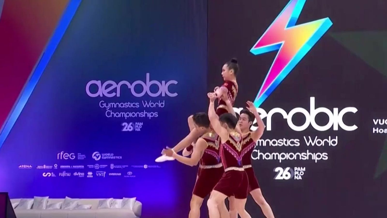 Cuatro gimnastas realizan una pirámide humana en un escenario con pantallas LED que muestran "aerobic Gymnastics World Championships".