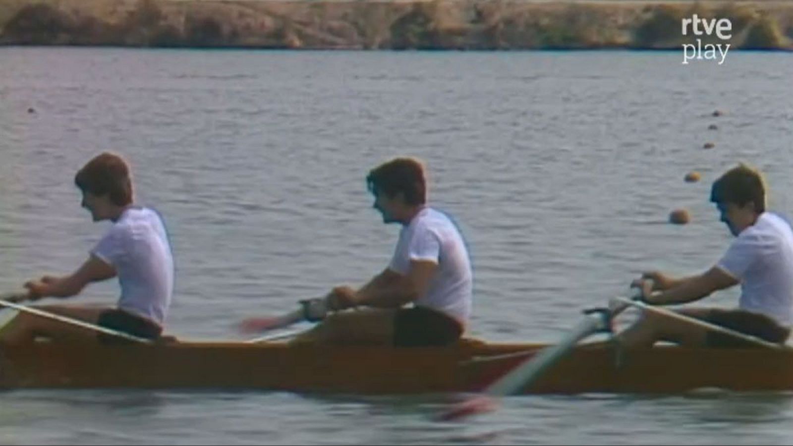 Tres hombres en un kayak K-3 compiten en aguas tranquilas, con boyas naranjas marcando el carril. Logo 'rtve play' en la esquina superior derecha.