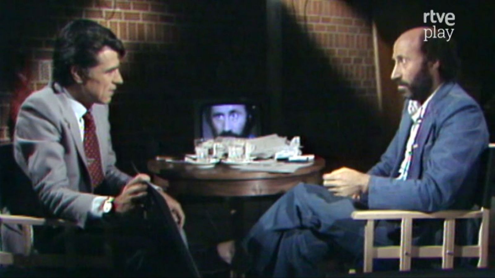 Jesús Hermida y Peridis en una entrevista de "rtve play". Ambos sentados frente a frente en un estudio con pared de ladrillo visto, mesa con vasos, papeles y un monitor que muestra un primer plano de Peridis.