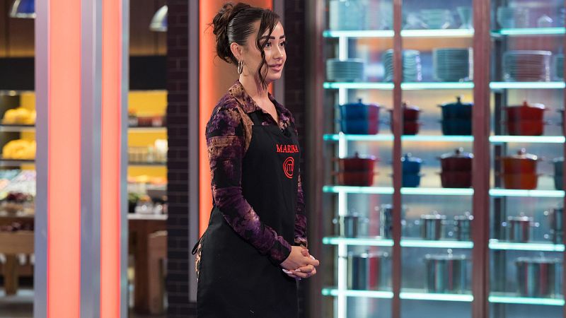 Marina Rivers en el set de "MasterChef Celebrity Legends" con delantal negro, camisa estampada y su nombre visible. Cocina profesional de fondo con estanterías iluminadas.