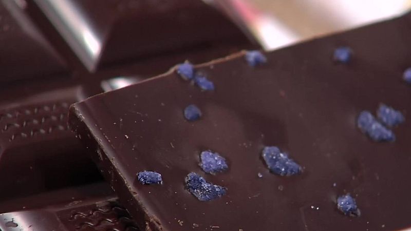 Chocolate oscuro con cristales azul violáceo esparcidos sobre su superficie lisa y brillante. Otros trozos de chocolate desenfocados al fondo.