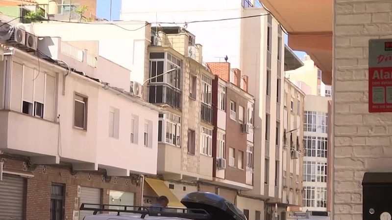 Calle urbana soleada con edificios, furgoneta blanca y cartel de "Alarma Securitas Direct" en pared de ladrillo blanco.
