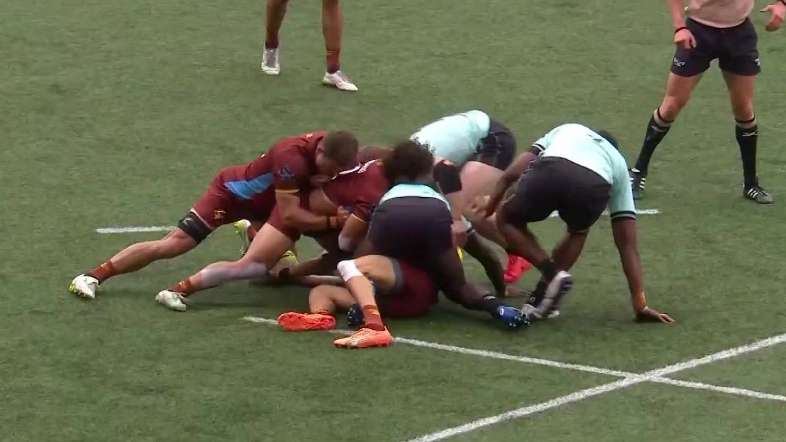 Jugadores de rugby en un ruck o maul, luchando por el balón en un campo verde. Equipos con uniformes granates y azules.