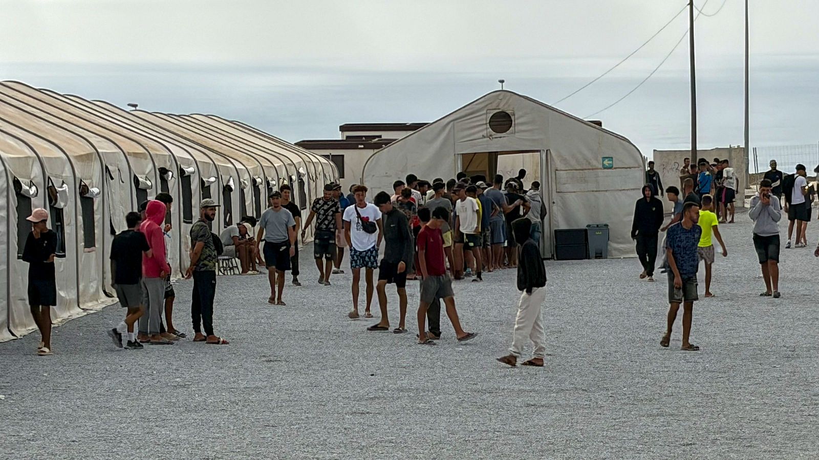 Vista amplia de un campamento con tiendas de campaña y un grupo de personas esperando en la entrada de una tienda central. Cielo cubierto y terreno de grava.