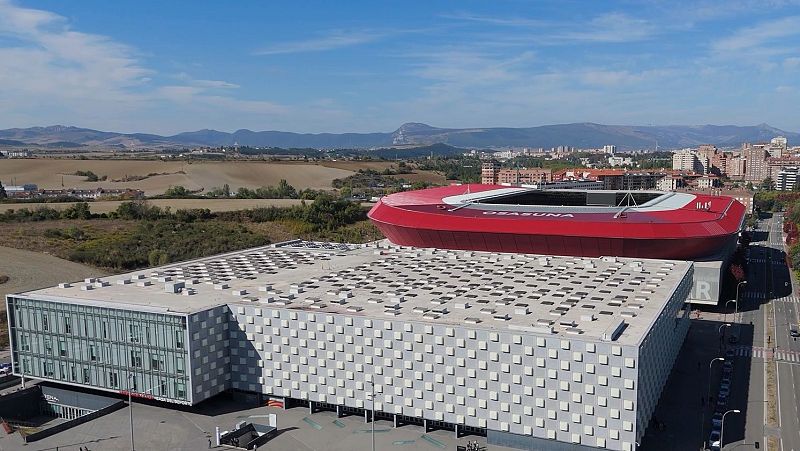 Vista aérea de Pamplona: Navarra Arena y Estadio El Sadar con "OSASUNA" visible. Entorno urbano y rural con montañas al fondo.