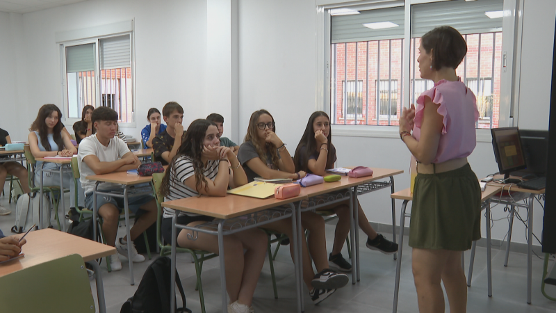 Profesora imparte clase a estudiantes en aula moderna. Se aprecian pupitres, materiales escolares y ventanas al fondo. Imagen de inicio de bachillerato.