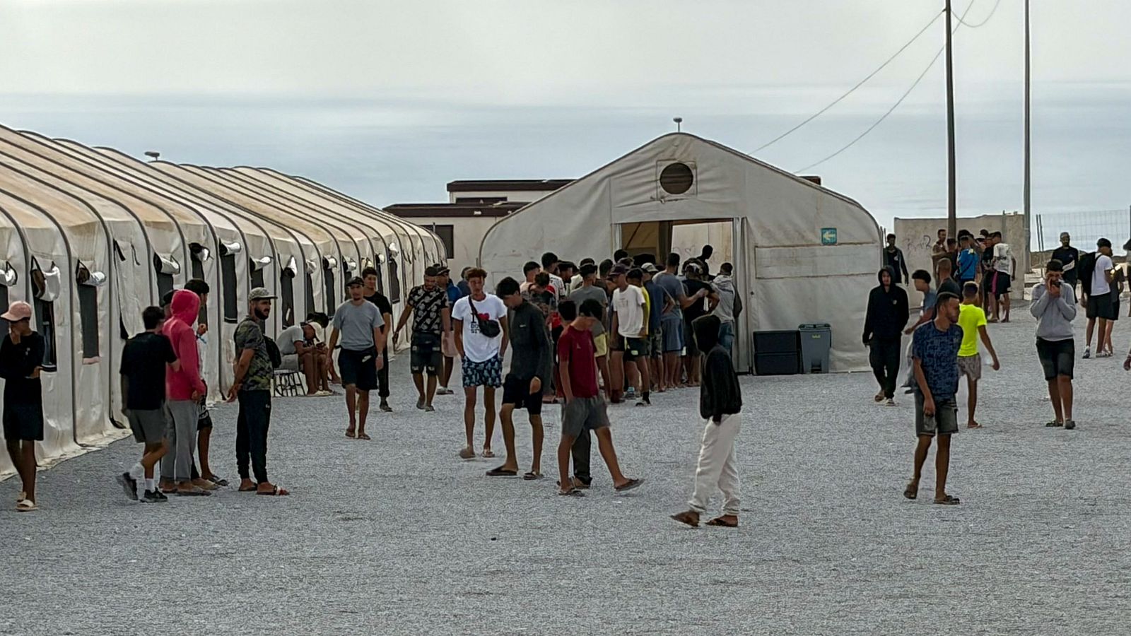 Campamento al aire libre con tiendas modulares y una tienda central grande, bajo cielo nublado. Numerosas personas, mayormente hombres jóvenes, transitan y se congregan en el área con suelo de grava.