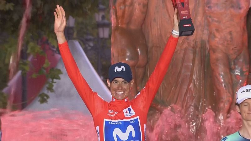 Ciclista Enric Mas celebra victoria con trofeo "LA VUELTA", luciendo maillot "MOVISTAR" y frente a fuente rosa.