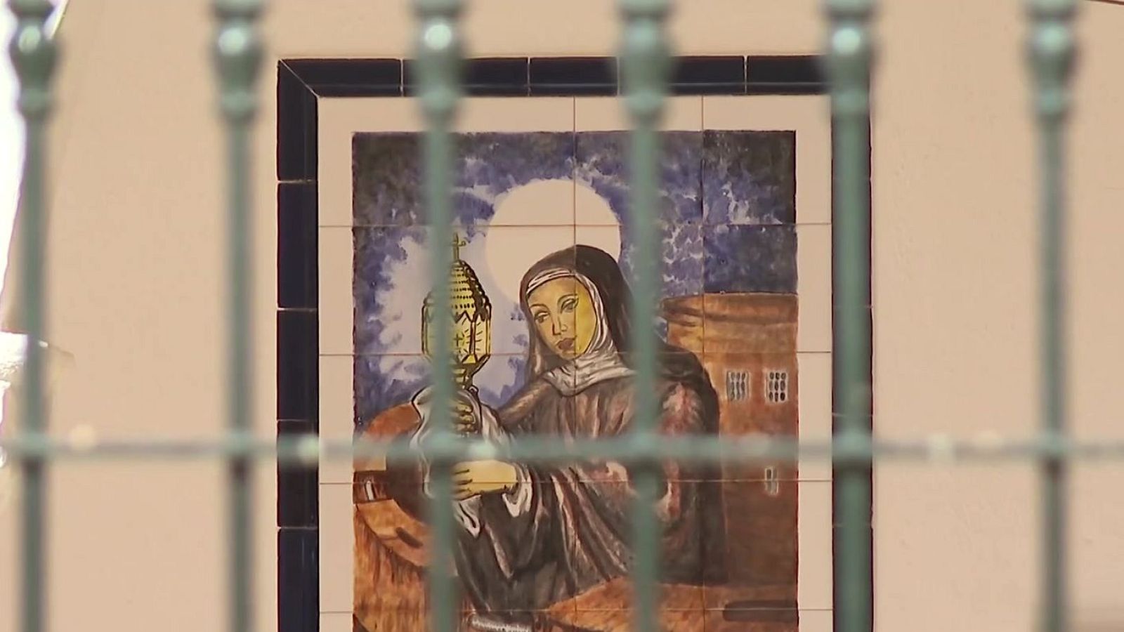 Mural de azulejos con figura religiosa femenina, halo y objeto sagrado, visto a través de una reja verde desenfocada.