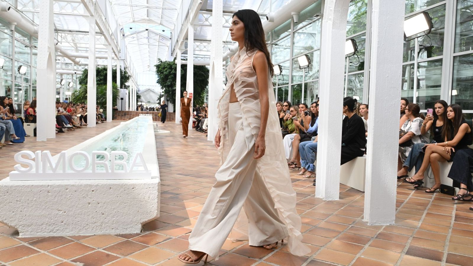 Desfile de moda en un espacio acristalado con letrero "SIMORRA". Modelo con conjunto crema camina por pasarela de terracota, espectadores observan.