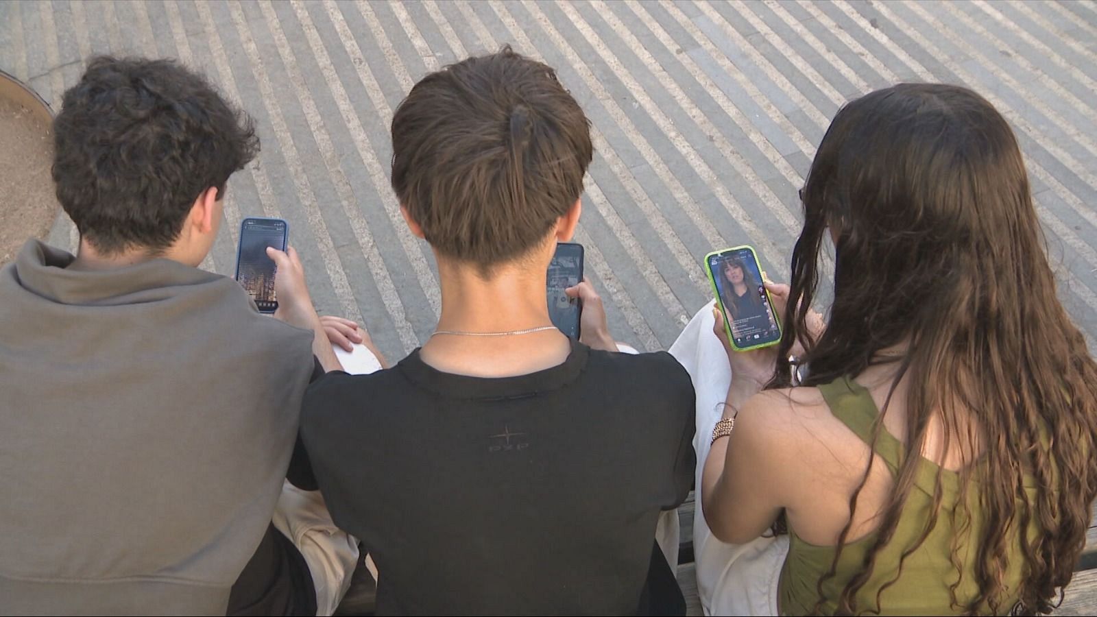 Tres jóvenes concentrados en sus teléfonos móviles en un espacio exterior con suelo de rayas diagonales. Se aprecian detalles en su vestimenta y accesorios, pero sus rostros no son visibles.